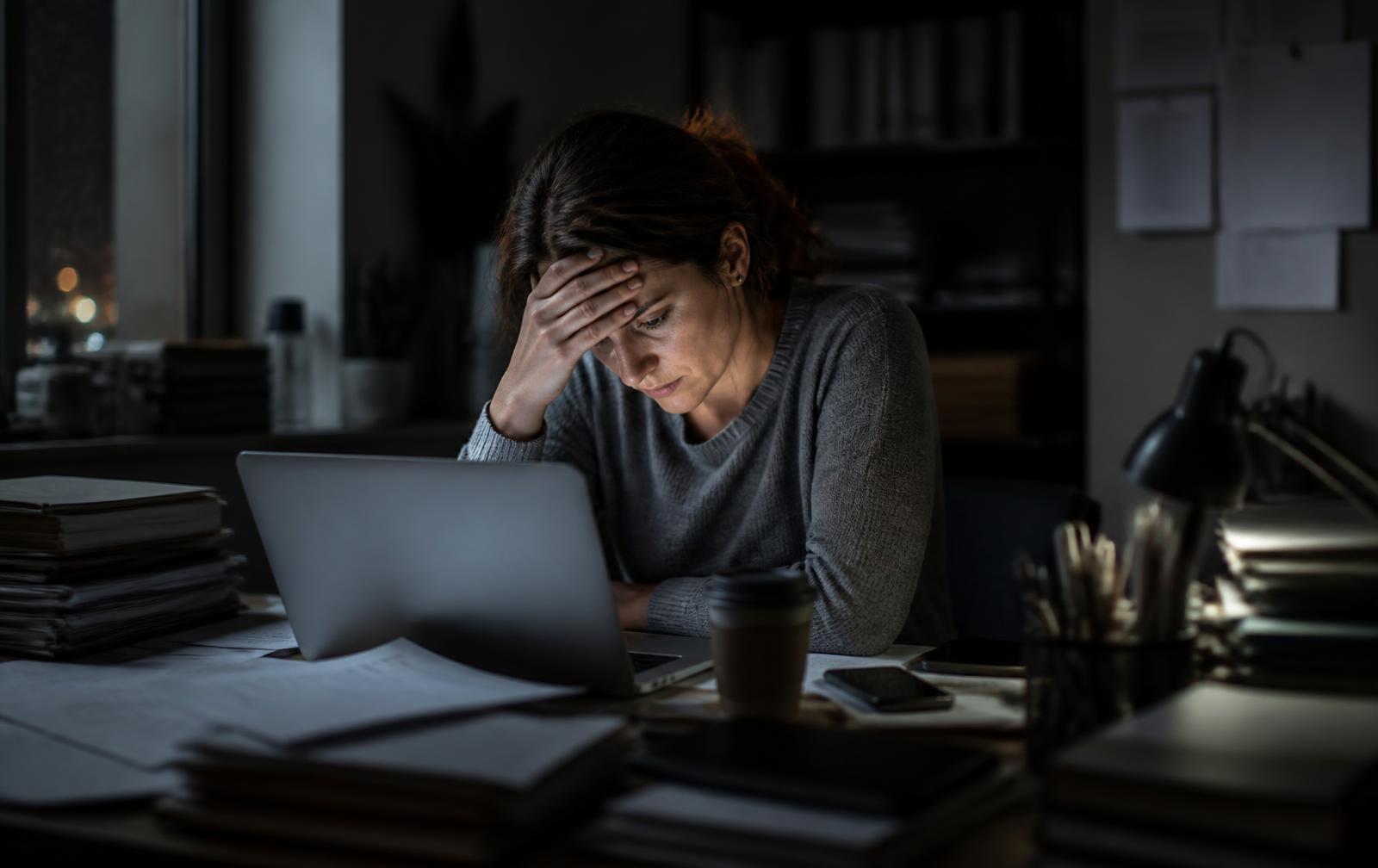 A person sitting at a cluttered desk late at night, illuminated by a laptop screen, with their hand pressed to their forehead in a gesture of stress.
